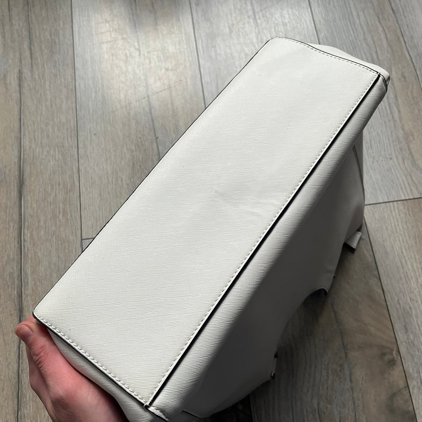 Steve Madden Removable Clutch White Handbag is a white leather tote featuring silver-tone hardware, including a front plaque and decorative buttons. The bag has two top handles and includes a removable inner clutch with silver Steve Madden branding. Designed for versatility, the clutch can be used separately or inside the main handbag.