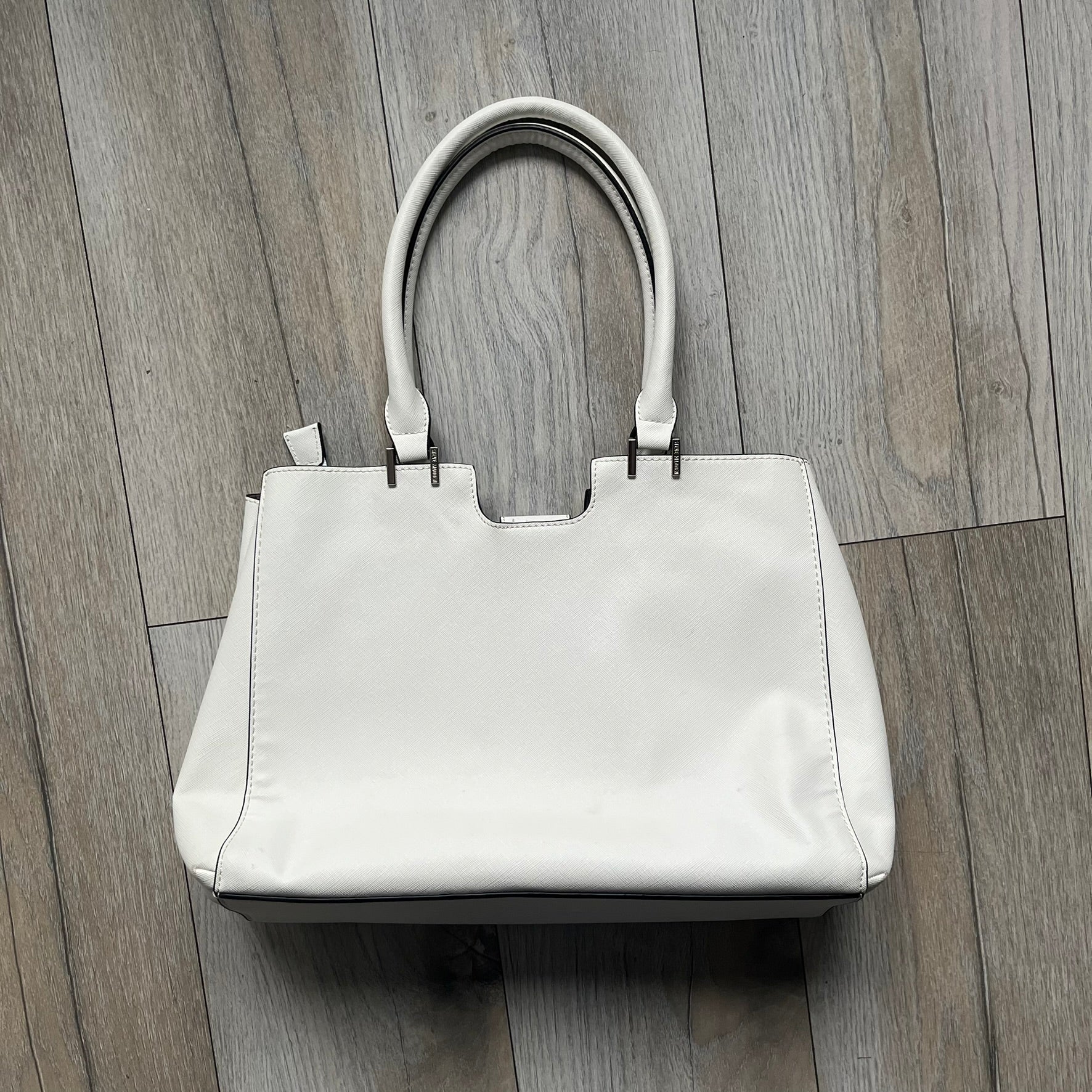 Steve Madden Removable Clutch White Handbag is a white leather tote featuring silver-tone hardware, including a front plaque and decorative buttons. The bag has two top handles and includes a removable inner clutch with silver Steve Madden branding. Designed for versatility, the clutch can be used separately or inside the main handbag.