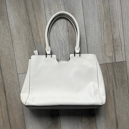 Steve Madden Removable Clutch White Handbag is a white leather tote featuring silver-tone hardware, including a front plaque and decorative buttons. The bag has two top handles and includes a removable inner clutch with silver Steve Madden branding. Designed for versatility, the clutch can be used separately or inside the main handbag.