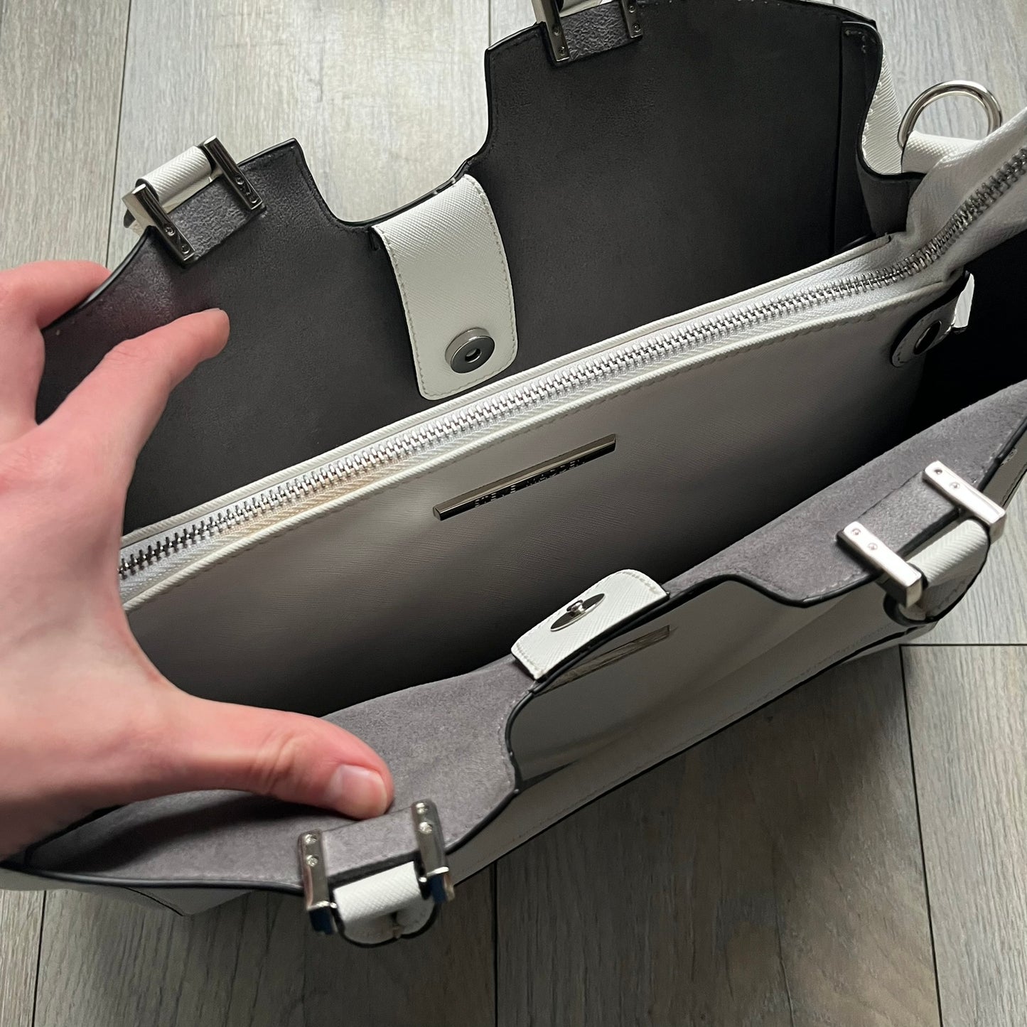 Steve Madden Removable Clutch White Handbag is a white leather tote featuring silver-tone hardware, including a front plaque and decorative buttons. The bag has two top handles and includes a removable inner clutch with silver Steve Madden branding. Designed for versatility, the clutch can be used separately or inside the main handbag.