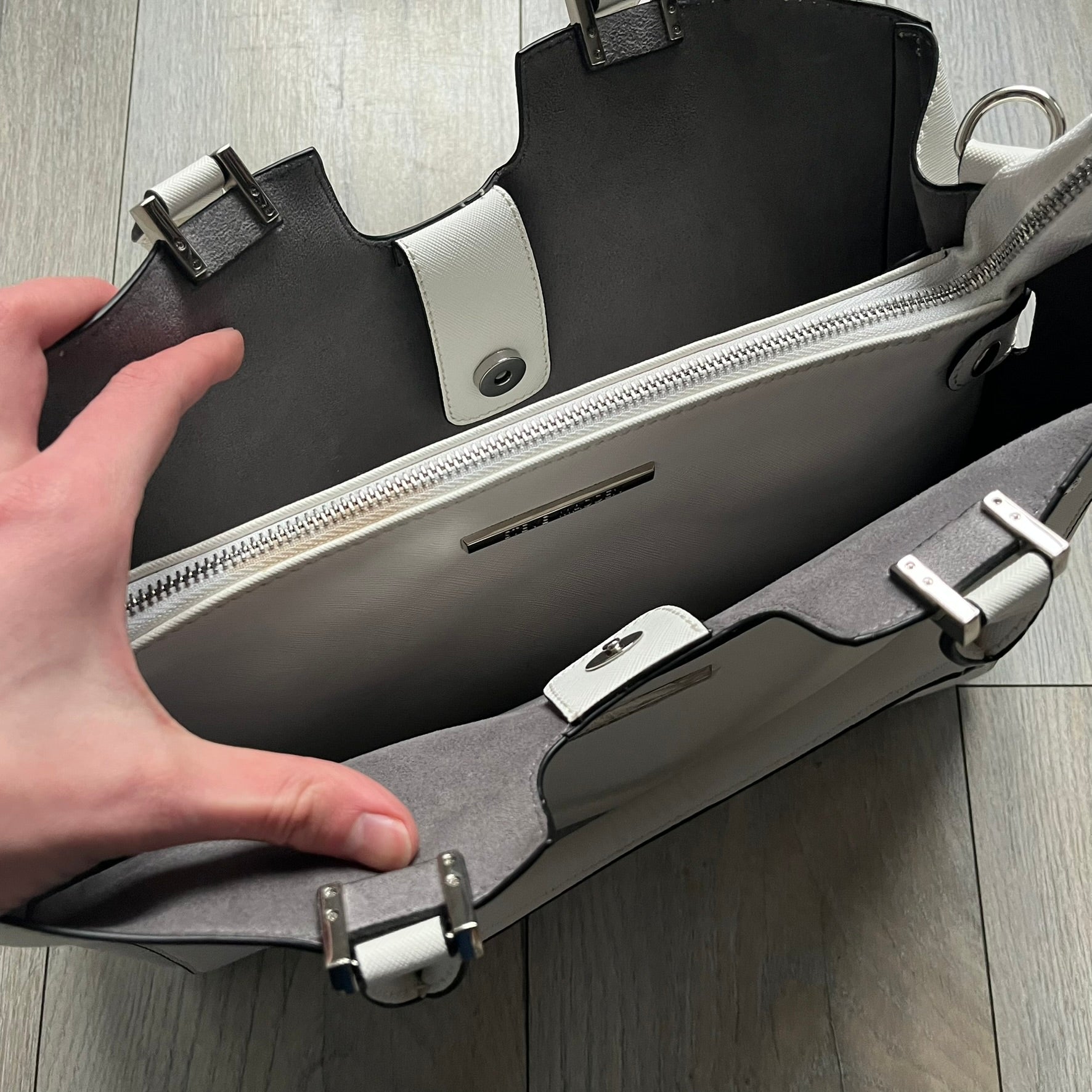 Steve Madden Removable Clutch White Handbag is a white leather tote featuring silver-tone hardware, including a front plaque and decorative buttons. The bag has two top handles and includes a removable inner clutch with silver Steve Madden branding. Designed for versatility, the clutch can be used separately or inside the main handbag.