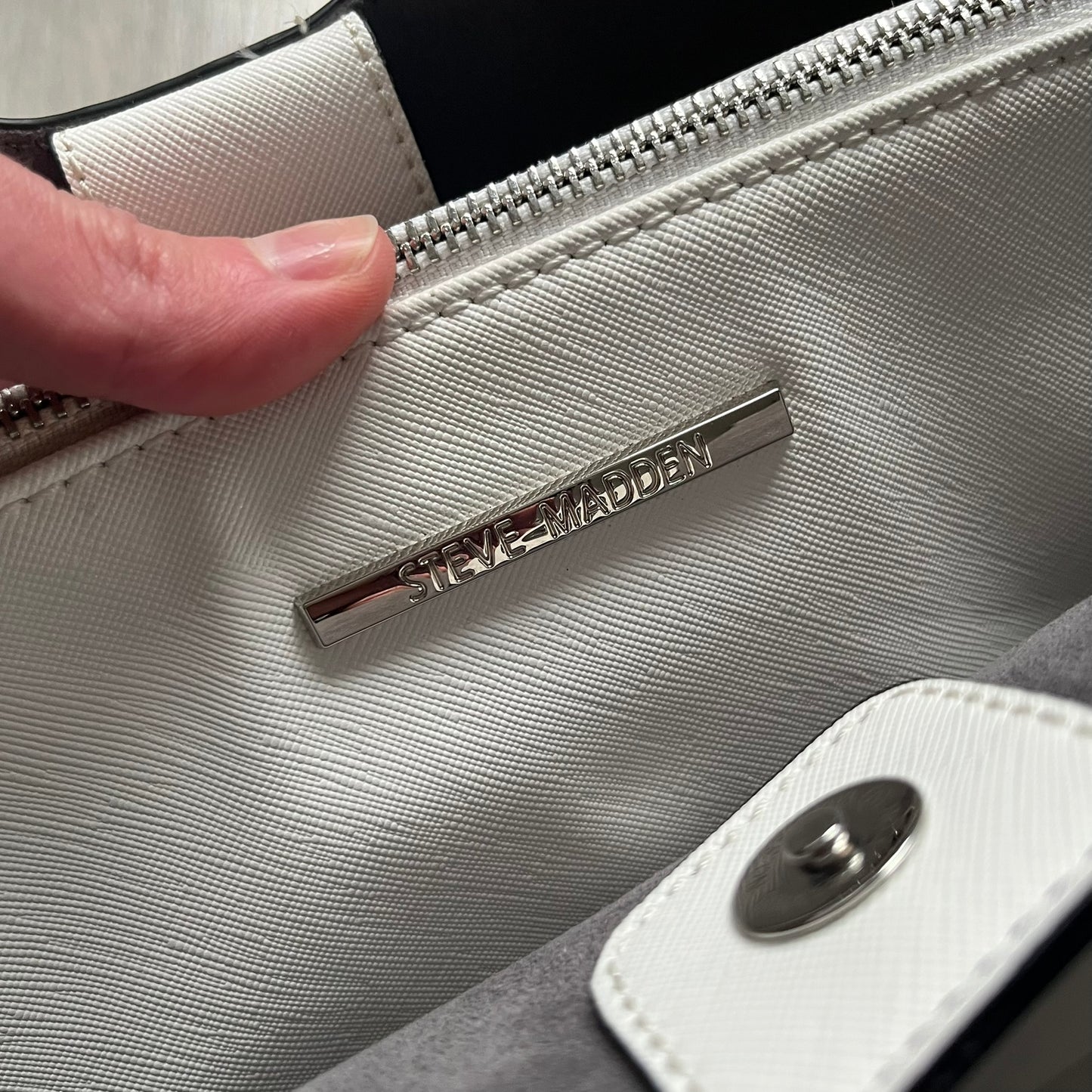 Steve Madden Removable Clutch White Handbag is a white leather tote featuring silver-tone hardware, including a front plaque and decorative buttons. The bag has two top handles and includes a removable inner clutch with silver Steve Madden branding. Designed for versatility, the clutch can be used separately or inside the main handbag.