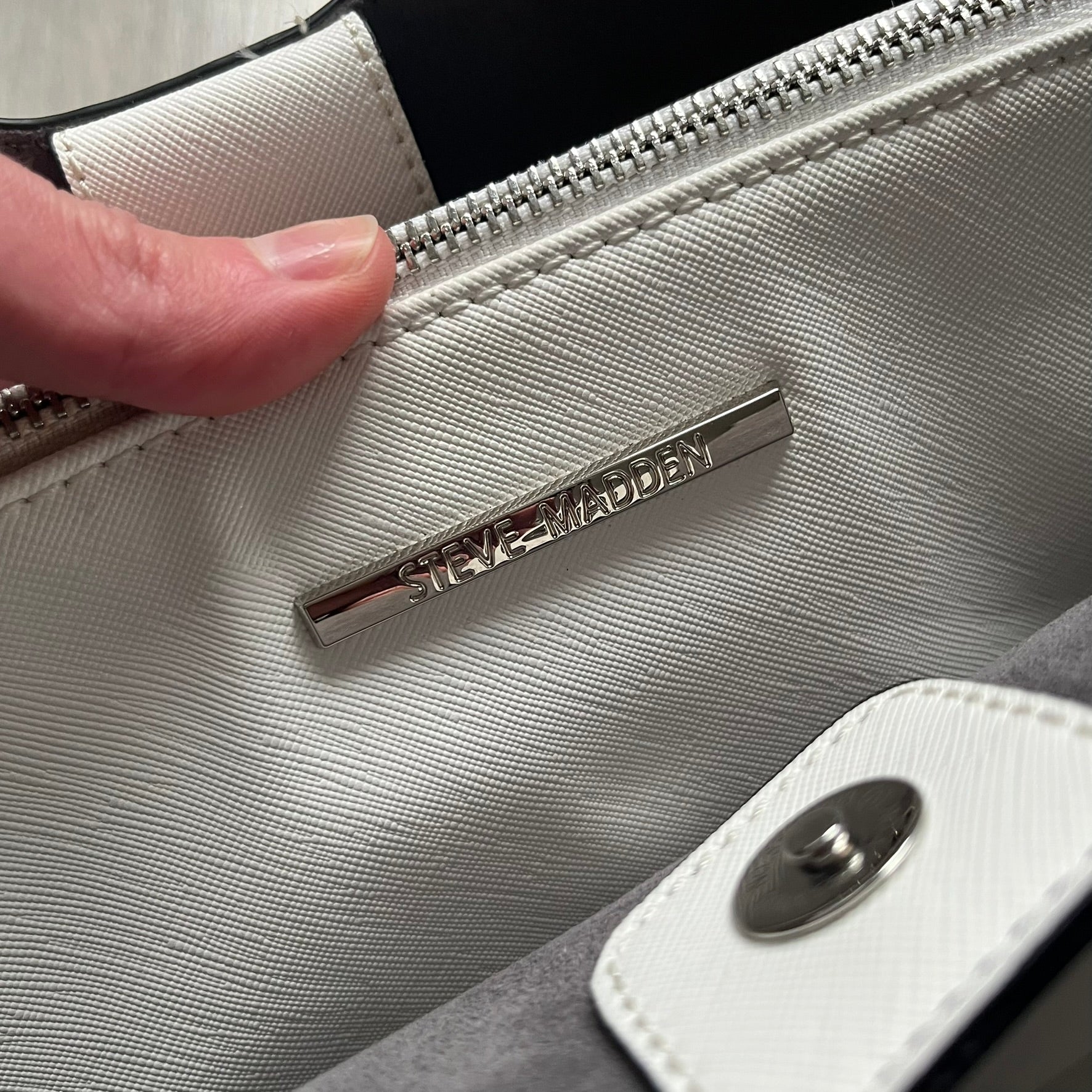 Steve Madden Removable Clutch White Handbag is a white leather tote featuring silver-tone hardware, including a front plaque and decorative buttons. The bag has two top handles and includes a removable inner clutch with silver Steve Madden branding. Designed for versatility, the clutch can be used separately or inside the main handbag.