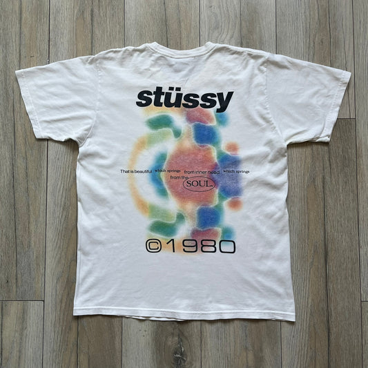 Stussy Soul 1980 White Tee is a white short-sleeve T-shirt released as part of a spring drop, made from soft cotton with a relaxed fit and ribbed collar. The front features a small Stussy logo print on the chest, while the back includes black Stussy text, a large multicolor central graphic, and “Copyright 1980” text at the bottom.