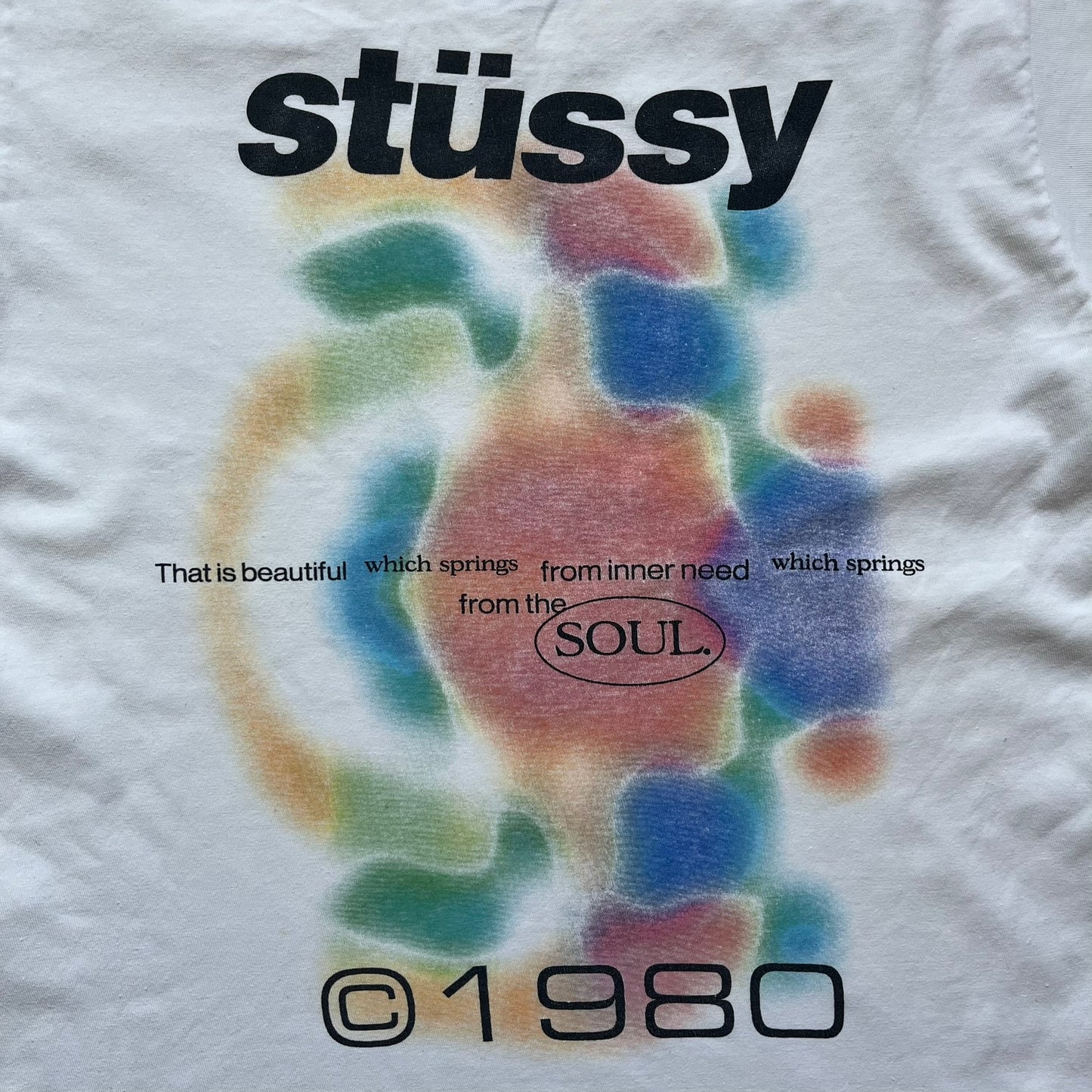 Stussy Soul 1980 White Tee is a white short-sleeve T-shirt released as part of a spring drop, made from soft cotton with a relaxed fit and ribbed collar. The front features a small Stussy logo print on the chest, while the back includes black Stussy text, a large multicolor central graphic, and “Copyright 1980” text at the bottom.