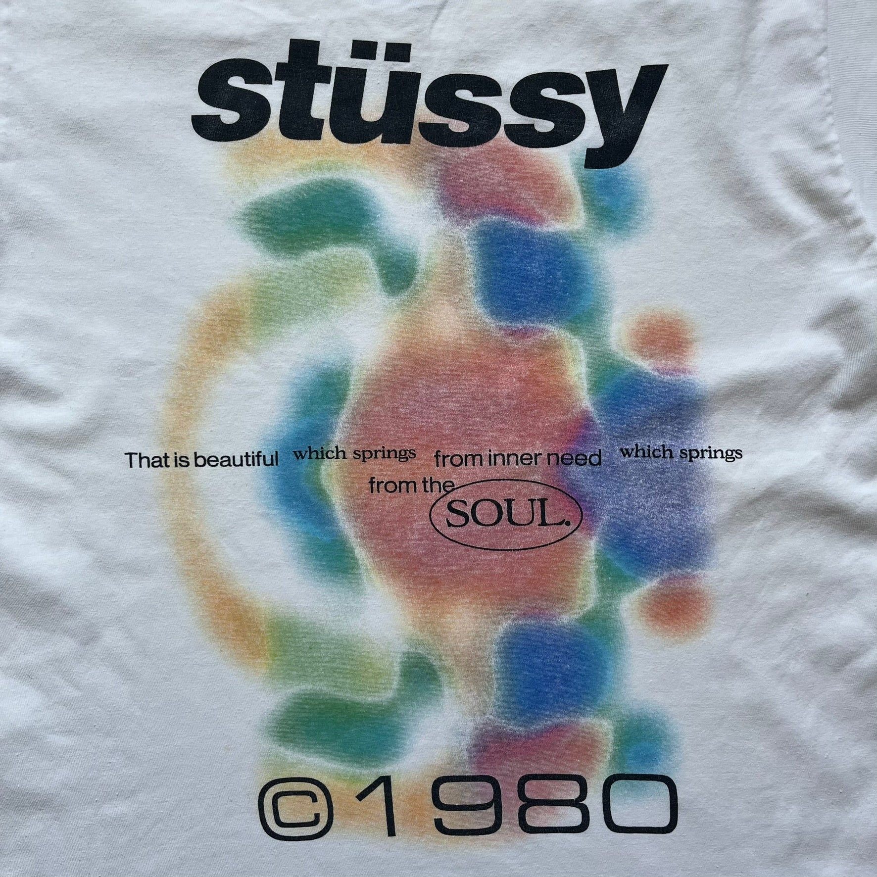 Stussy Soul 1980 White Tee is a white short-sleeve T-shirt released as part of a spring drop, made from soft cotton with a relaxed fit and ribbed collar. The front features a small Stussy logo print on the chest, while the back includes black Stussy text, a large multicolor central graphic, and “Copyright 1980” text at the bottom.
