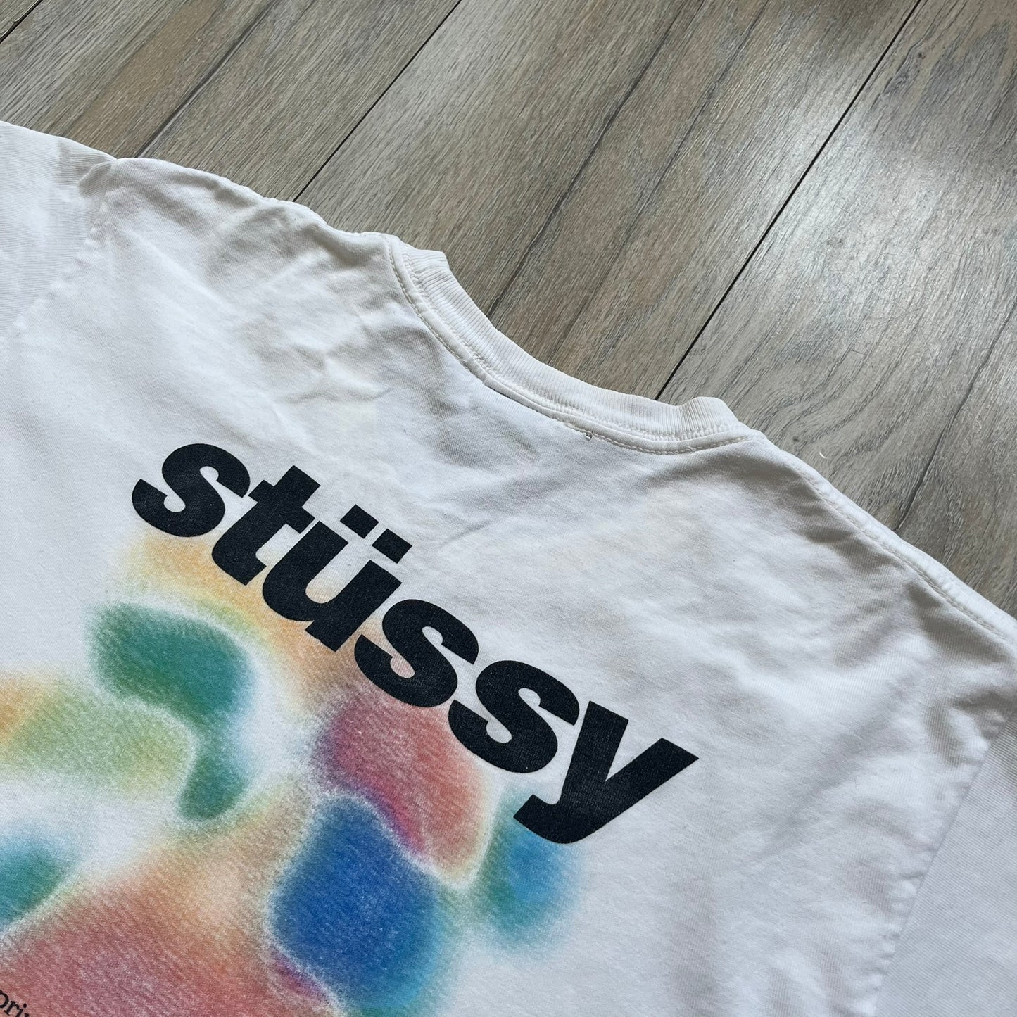 Stussy Soul 1980 White Tee is a white short-sleeve T-shirt released as part of a spring drop, made from soft cotton with a relaxed fit and ribbed collar. The front features a small Stussy logo print on the chest, while the back includes black Stussy text, a large multicolor central graphic, and “Copyright 1980” text at the bottom.