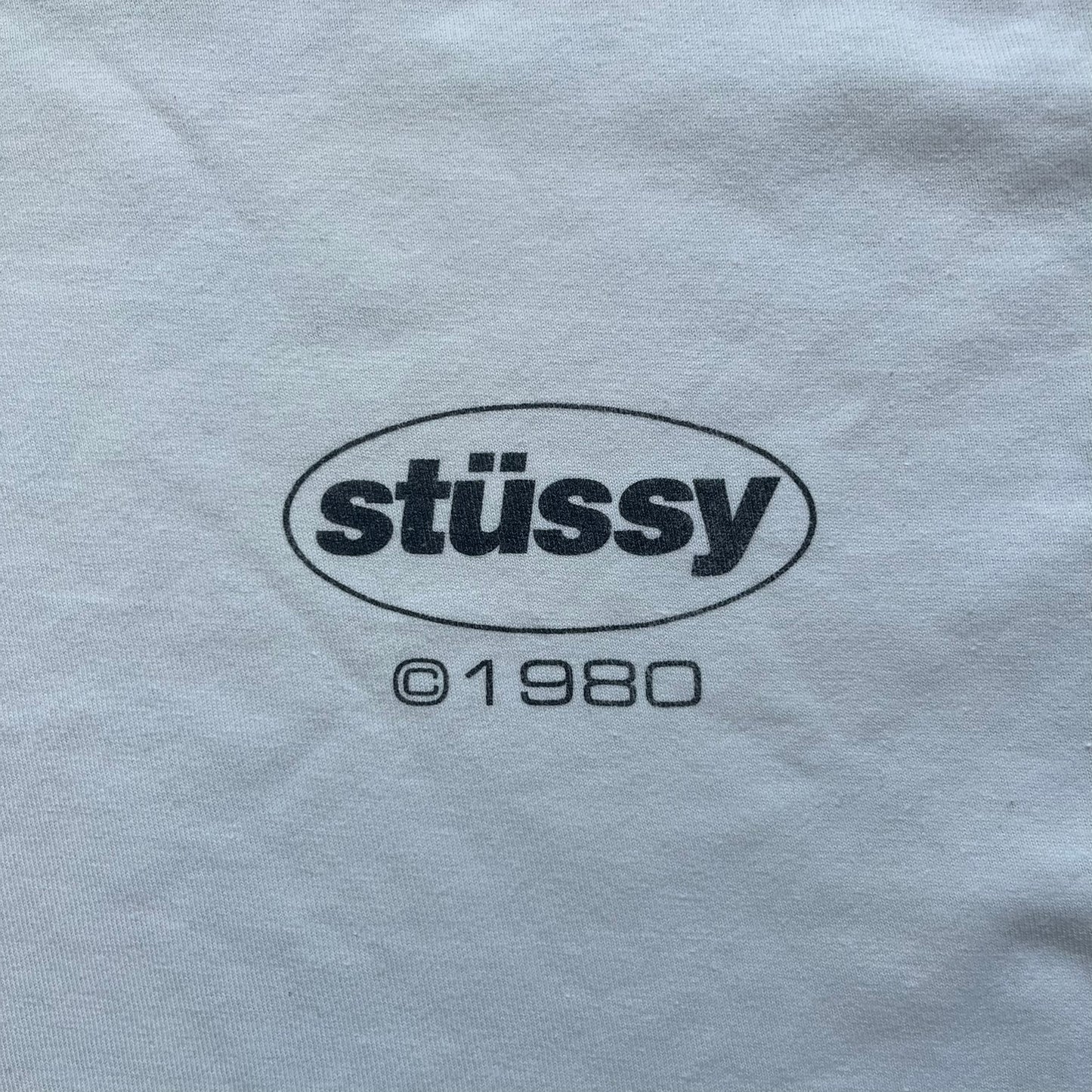 Stussy Soul 1980 White Tee is a white short-sleeve T-shirt released as part of a spring drop, made from soft cotton with a relaxed fit and ribbed collar. The front features a small Stussy logo print on the chest, while the back includes black Stussy text, a large multicolor central graphic, and “Copyright 1980” text at the bottom.