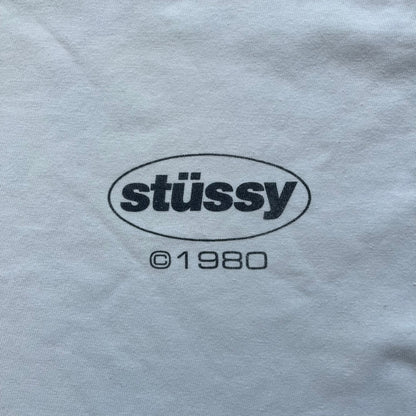 Stussy Soul 1980 White Tee is a white short-sleeve T-shirt released as part of a spring drop, made from soft cotton with a relaxed fit and ribbed collar. The front features a small Stussy logo print on the chest, while the back includes black Stussy text, a large multicolor central graphic, and “Copyright 1980” text at the bottom.