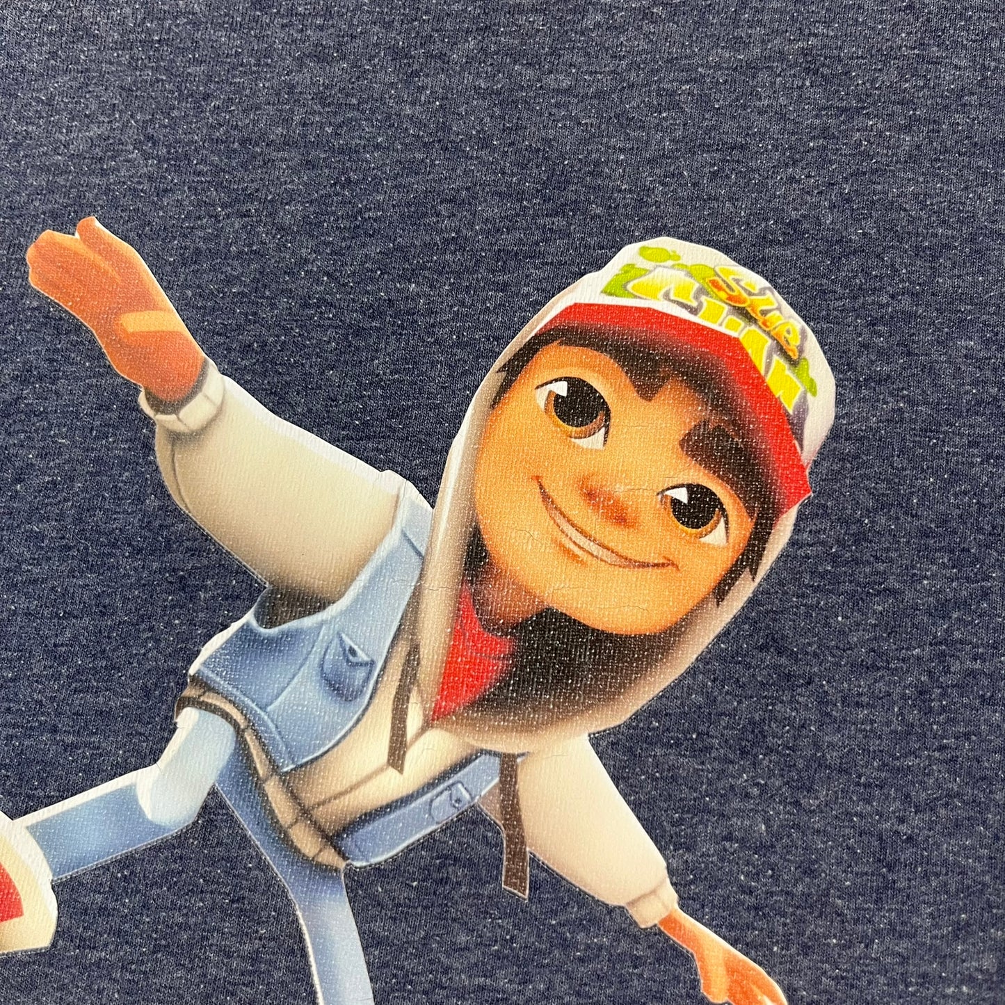 Subway Surfers 2010s Jake Tee is a grey T-shirt made on a Fruit of the Loom blank. The front features Jake, the main character from Subway Surfers, riding his skateboard, with the pursuing guard dog biting the end of the board. The design is printed in full color on the front.
