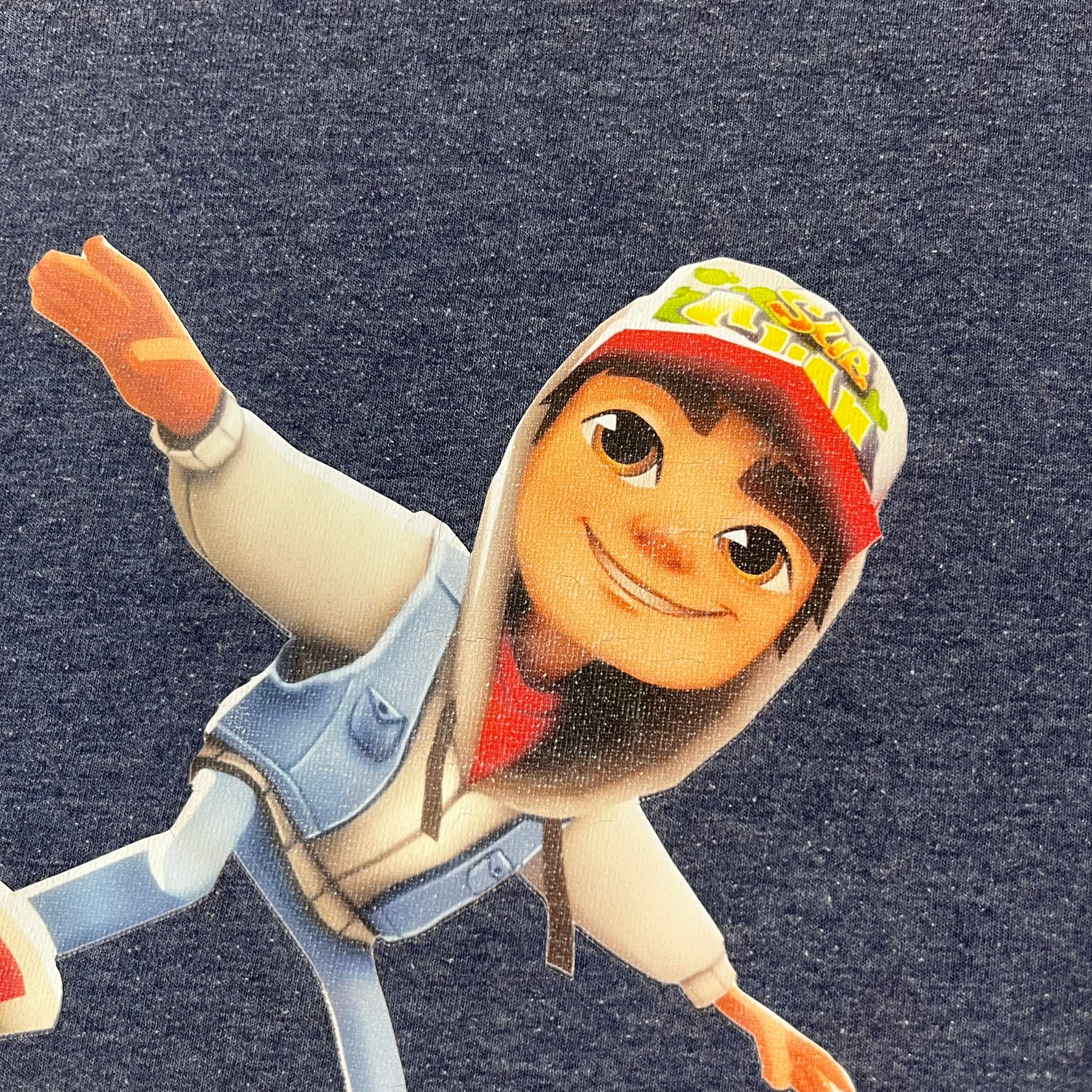 Subway Surfers 2010s Jake Tee is a grey T-shirt made on a Fruit of the Loom blank. The front features Jake, the main character from Subway Surfers, riding his skateboard, with the pursuing guard dog biting the end of the board. The design is printed in full color on the front.
