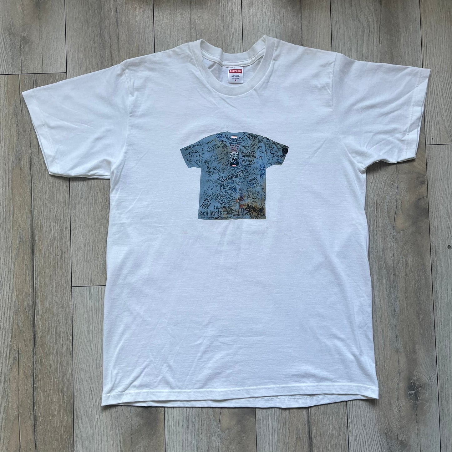 Supreme 30th Anniversary SS24 First Tee is a white T-shirt released on April 25, 2024, as part of the SS24 season. The front features a small OG Taxi Driver Supreme shirt graphic in blue with graffiti-style detailing. Below the back collar, a small gray tag displays the Supreme 30th Anniversary logo.