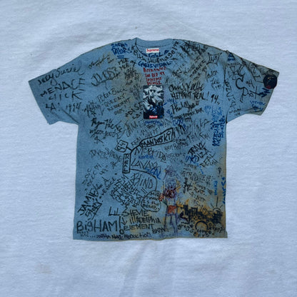 Supreme 30th Anniversary SS24 First Tee is a white T-shirt released on April 25, 2024, as part of the SS24 season. The front features a small OG Taxi Driver Supreme shirt graphic in blue with graffiti-style detailing. Below the back collar, a small gray tag displays the Supreme 30th Anniversary logo.