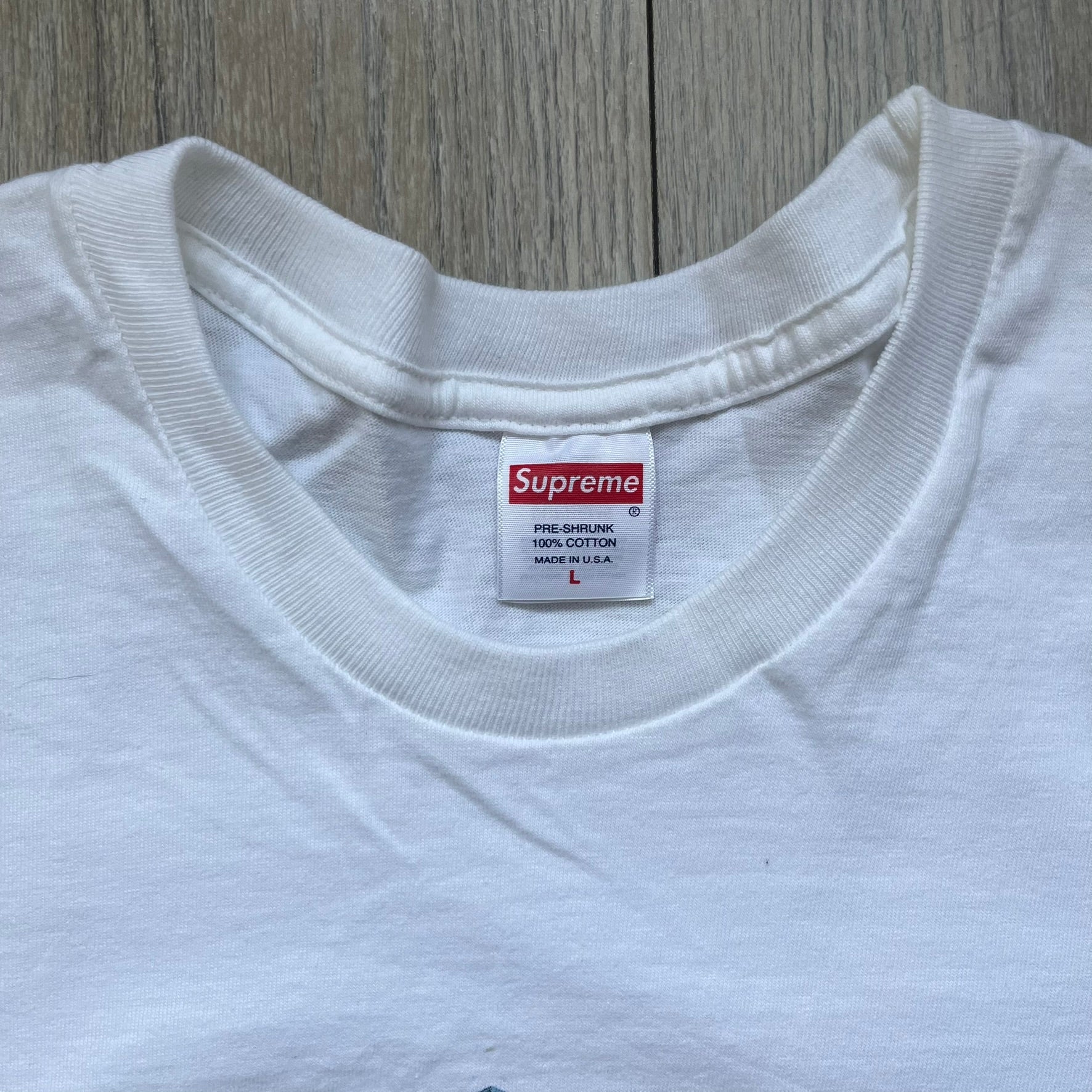 Supreme 30th Anniversary SS24 First Tee is a white T-shirt released on April 25, 2024, as part of the SS24 season. The front features a small OG Taxi Driver Supreme shirt graphic in blue with graffiti-style detailing. Below the back collar, a small gray tag displays the Supreme 30th Anniversary logo.
