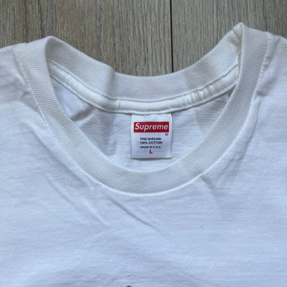 Supreme 30th Anniversary SS24 First Tee is a white T-shirt released on April 25, 2024, as part of the SS24 season. The front features a small OG Taxi Driver Supreme shirt graphic in blue with graffiti-style detailing. Below the back collar, a small gray tag displays the Supreme 30th Anniversary logo.