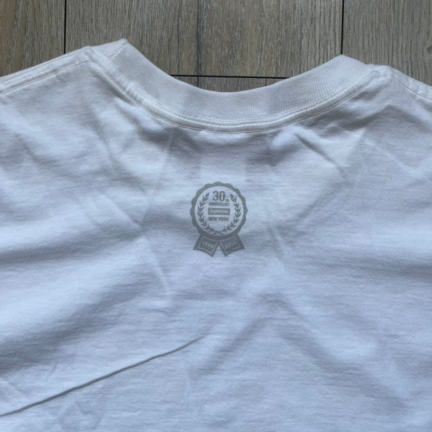 Supreme 30th Anniversary SS24 First Tee is a white T-shirt released on April 25, 2024, as part of the SS24 season. The front features a small OG Taxi Driver Supreme shirt graphic in blue with graffiti-style detailing. Below the back collar, a small gray tag displays the Supreme 30th Anniversary logo.