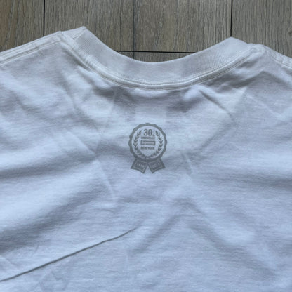 Supreme 30th Anniversary SS24 First Tee is a white T-shirt released on April 25, 2024, as part of the SS24 season. The front features a small OG Taxi Driver Supreme shirt graphic in blue with graffiti-style detailing. Below the back collar, a small gray tag displays the Supreme 30th Anniversary logo.