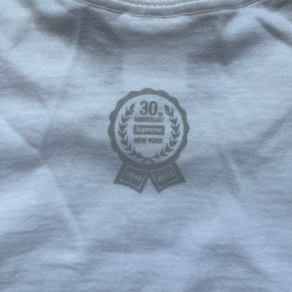 Supreme 30th Anniversary SS24 First Tee is a white T-shirt released on April 25, 2024, as part of the SS24 season. The front features a small OG Taxi Driver Supreme shirt graphic in blue with graffiti-style detailing. Below the back collar, a small gray tag displays the Supreme 30th Anniversary logo.