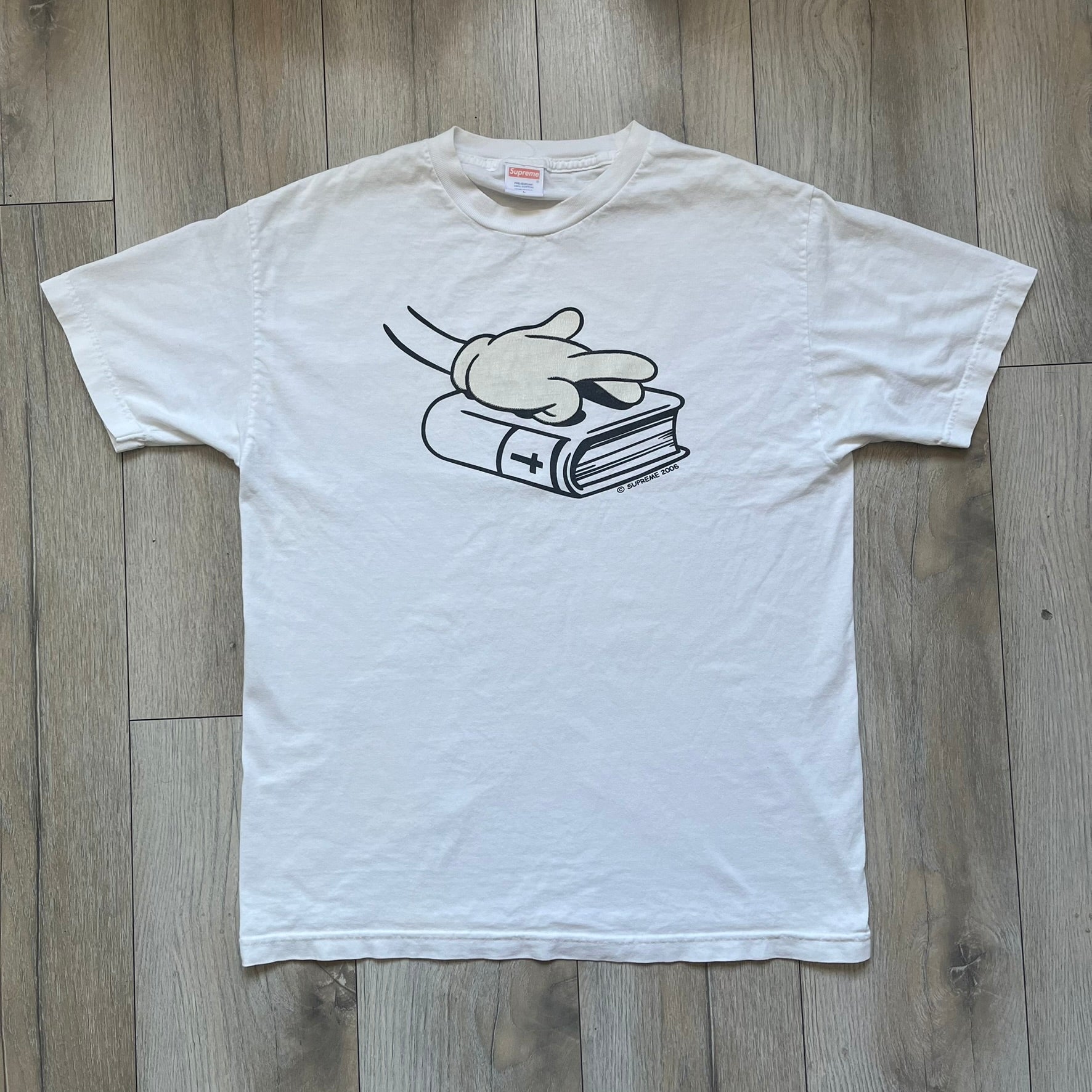 Supreme SS06 Oath White Tee is a white short-sleeve T-shirt from the Spring/Summer 2006 collection. The front features a black outline graphic of a Mickey Mouse-style hand placed on a cartoon Bible with a visible cross, with small “Supreme 2006” copyright text on the side. Super rare piece.