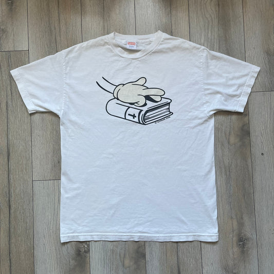 Supreme SS06 Oath White Tee is a white short-sleeve T-shirt from the Spring/Summer 2006 collection. The front features a black outline graphic of a Mickey Mouse-style hand placed on a cartoon Bible with a visible cross, with small “Supreme 2006” copyright text on the side. Super rare piece.
