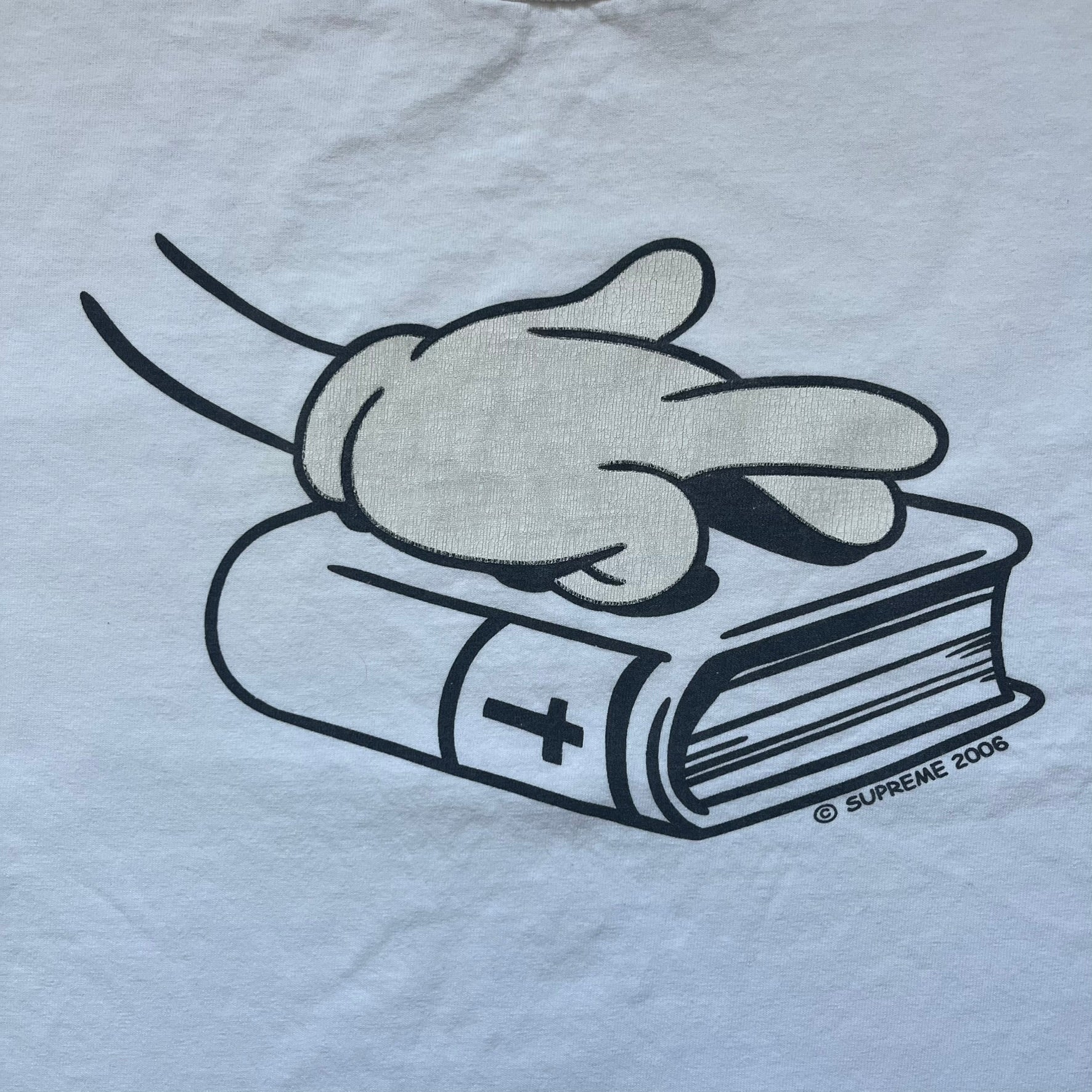 Supreme SS06 Oath White Tee is a white short-sleeve T-shirt from the Spring/Summer 2006 collection. The front features a black outline graphic of a Mickey Mouse-style hand placed on a cartoon Bible with a visible cross, with small “Supreme 2006” copyright text on the side. Super rare piece.