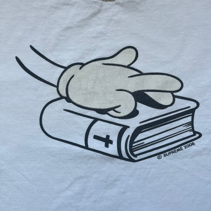 Supreme SS06 Oath White Tee is a white short-sleeve T-shirt from the Spring/Summer 2006 collection. The front features a black outline graphic of a Mickey Mouse-style hand placed on a cartoon Bible with a visible cross, with small “Supreme 2006” copyright text on the side. Super rare piece.