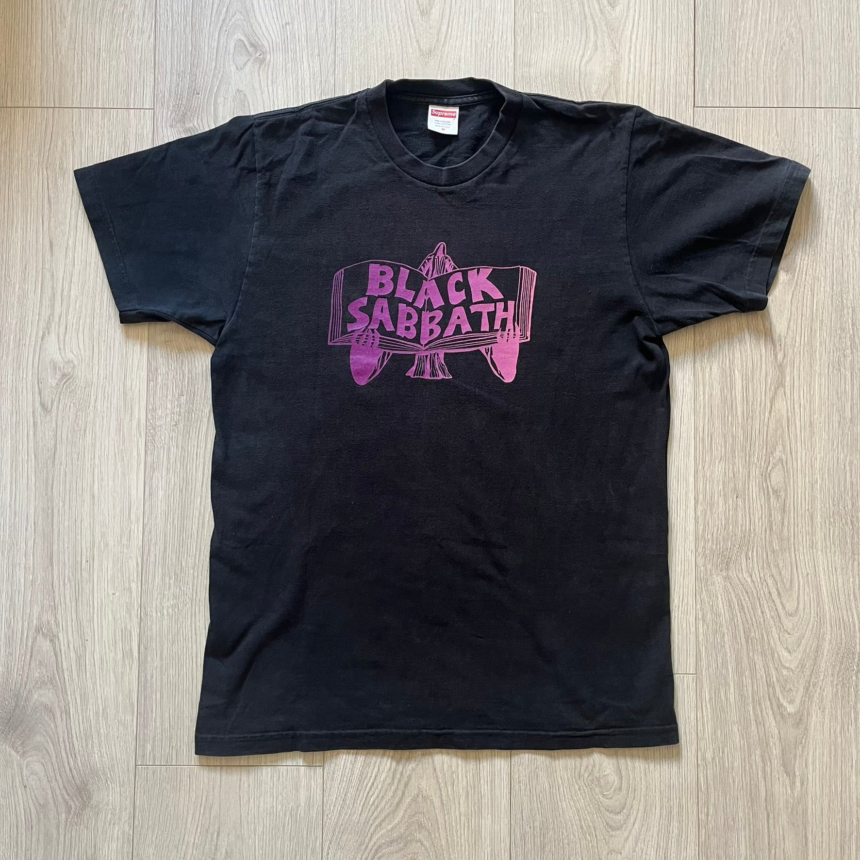 From Supreme’s SS16 Black Sabbath collaboration, the Tome Tee arrives in black with striking purple graphics. The front shows the band’s Children of the Grave artwork within a book, contrasted by a gothic Supreme logo on the back. A sought-after item bridging metal iconography and Supreme’s streetwear legacy.