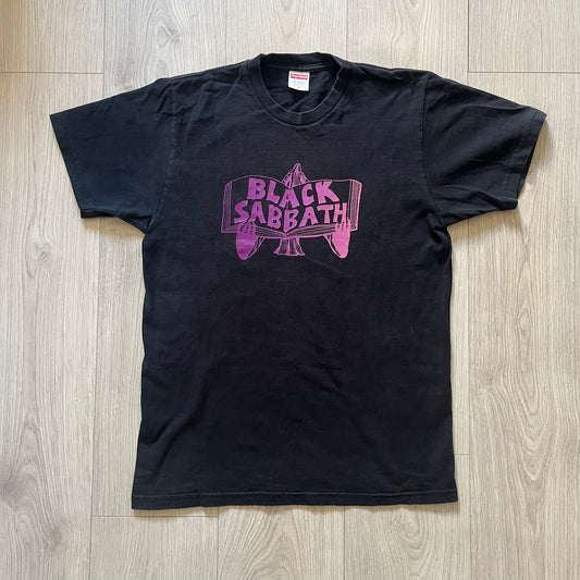 From Supreme’s SS16 Black Sabbath collaboration, the Tome Tee arrives in black with striking purple graphics. The front shows the band’s Children of the Grave artwork within a book, contrasted by a gothic Supreme logo on the back. A sought-after item bridging metal iconography and Supreme’s streetwear legacy.