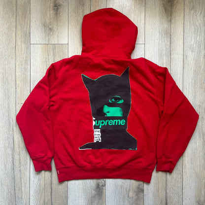 Supreme Catwoman FW23 Hooded Sweatshirt is a red pullover hoodie released on October 19, 2023. It features a drawstring hood, kangaroo pocket, and ribbed cuffs and hem. A small Supreme box logo is placed on the left chest in black.&nbsp;The back features a large Catwoman mask graphic combined with Supreme branding across the width of the design in black and green.
