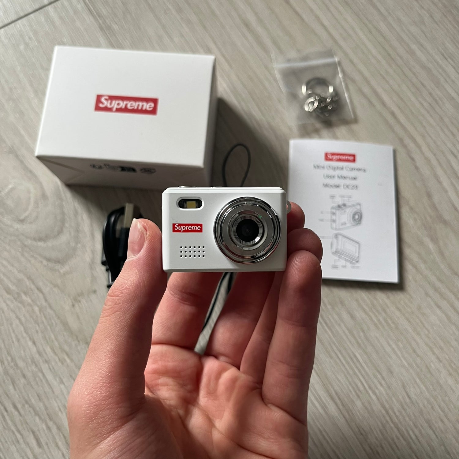 Supreme Digital Camera Keychain released for FW25 in white with a printed Supreme box logo on the front. This ultra-compact digital camera measures 1.92" x 1.14" x 1.1" and includes a 32GB TF card, USB-C cable, and built-in 1080p HD recording. When powered on, the screen displays the Supreme logo, with selectable interface colors in colors like: pink, green, or blue.