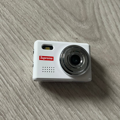 Supreme Digital Camera Keychain released for FW25 in white with a printed Supreme box logo on the front. This ultra-compact digital camera measures 1.92" x 1.14" x 1.1" and includes a 32GB TF card, USB-C cable, and built-in 1080p HD recording. When powered on, the screen displays the Supreme logo, with selectable interface colors in colors like: pink, green, or blue.