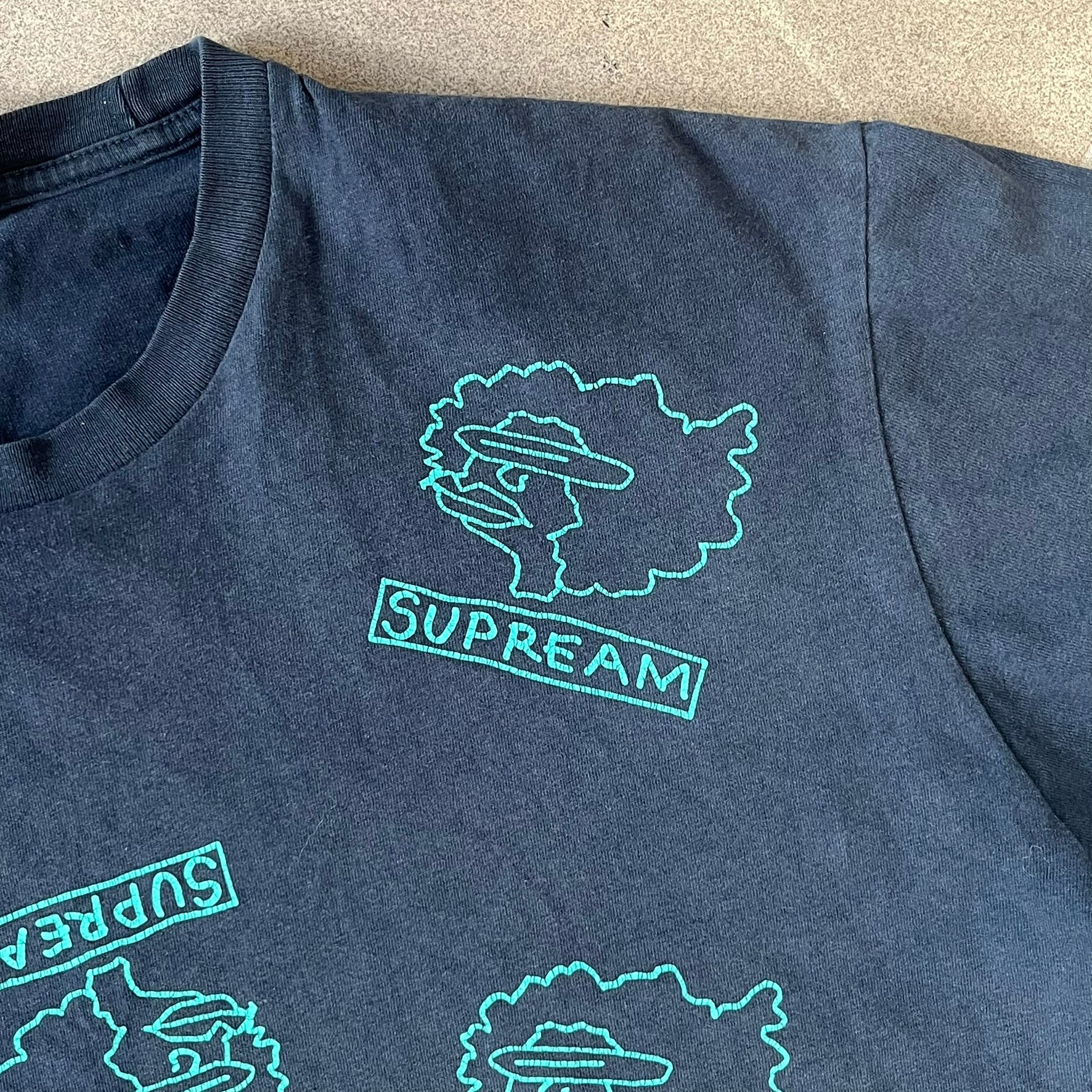 Supreme FW17 Gonz Heads Tee is a navy T-shirt from the Fall/Winter 2017 collection created in collaboration with Mark Gonzales (The Gonz). The tee features repeated green Gonz head graphics printed across both the front and back in different orientations. The shirt is made in the USA.
