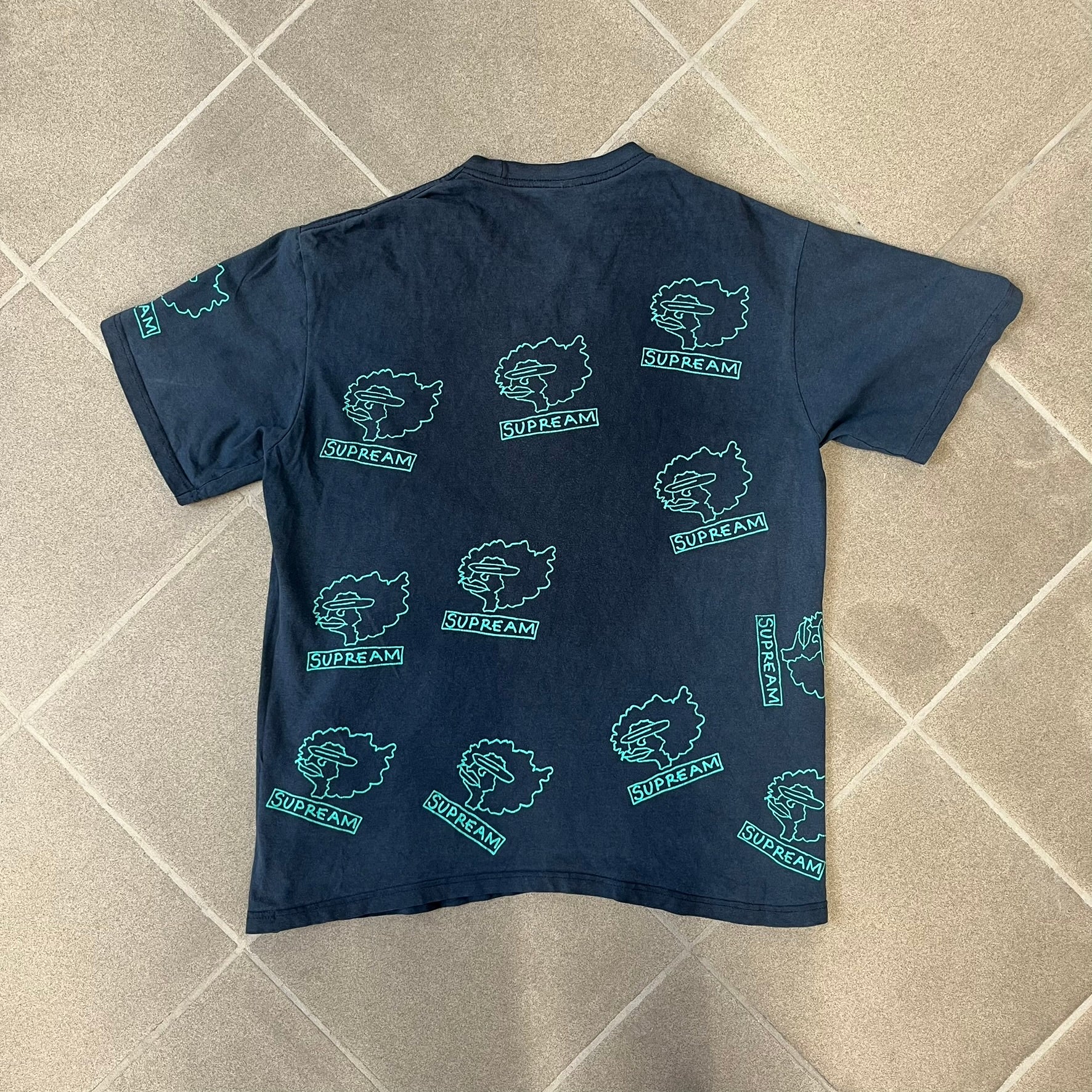 Supreme FW17 Gonz Heads Tee is a navy T-shirt from the Fall/Winter 2017 collection created in collaboration with Mark Gonzales (The Gonz). The tee features repeated green Gonz head graphics printed across both the front and back in different orientations. The shirt is made in the USA.
