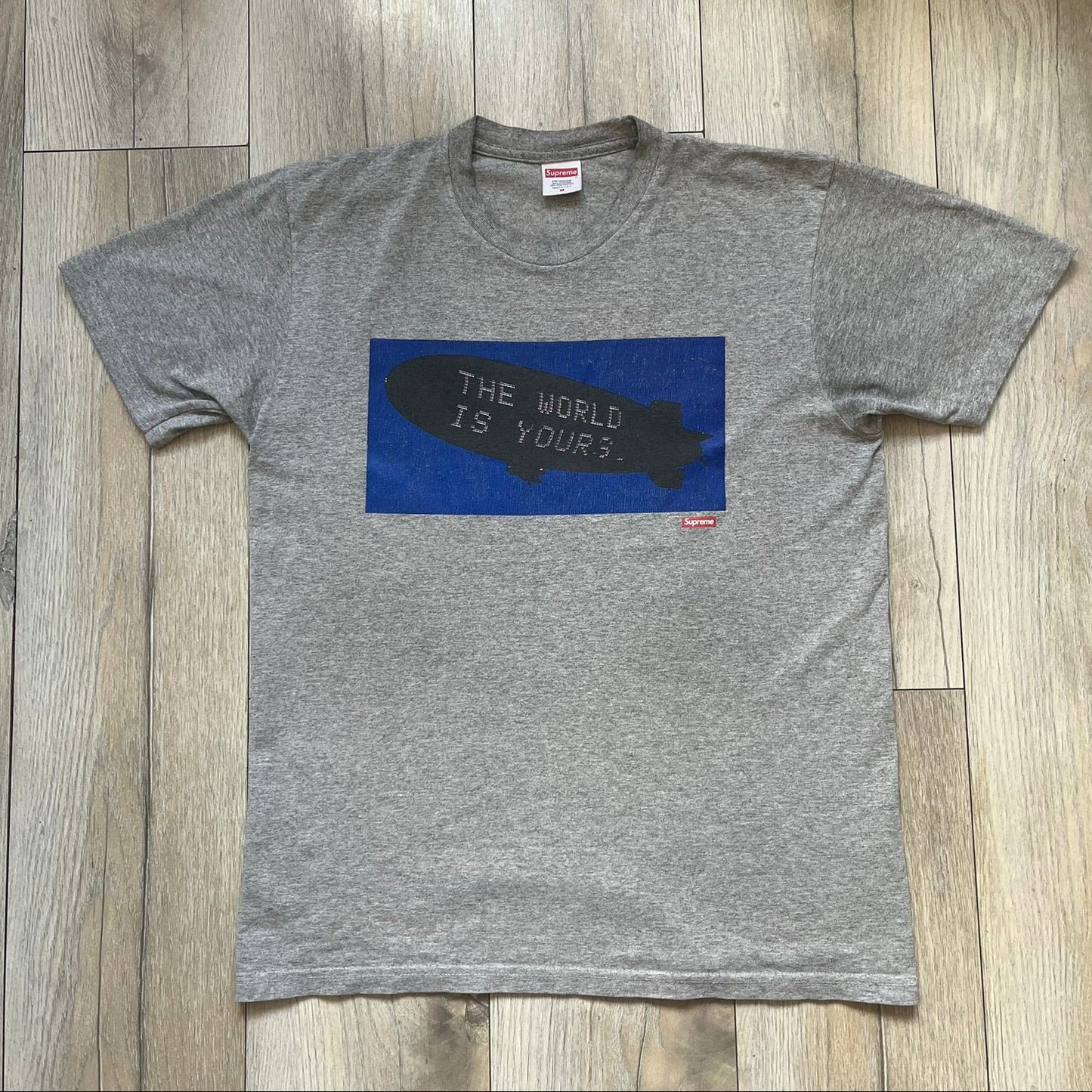 Supreme FW17 Scarface Blimp Gray Tee is a grey T-shirt from the Fall/Winter 2017 collection featuring a large front print from the film Scarface. The graphic shows the blimp scene with “The World Is Yours” text in black on a blue background, with a small red Supreme box logo printed below. Released in October 2017 as part of the Supreme x Scarface collaboration.