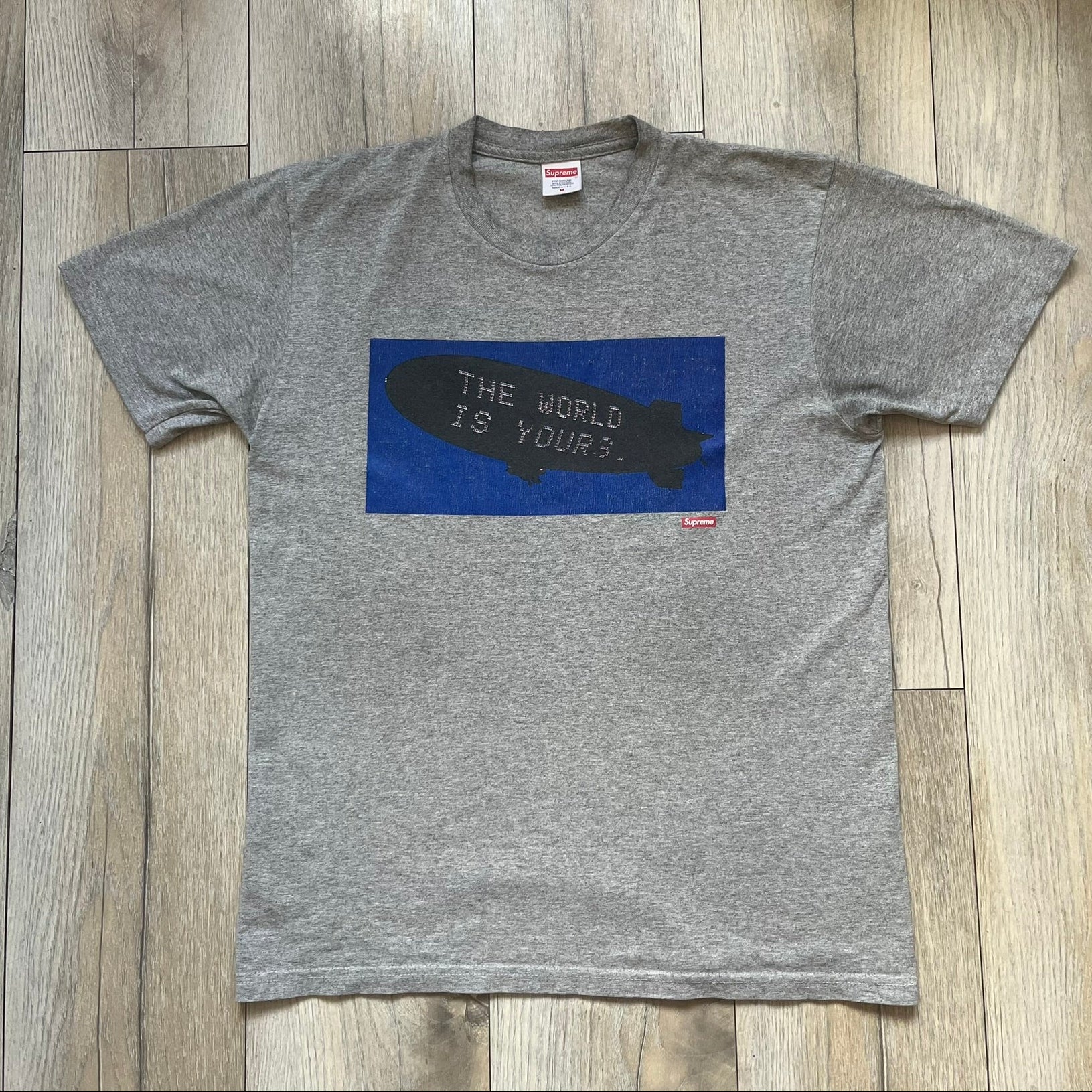 Supreme FW17 Scarface Blimp Gray Tee is a grey T-shirt from the Fall/Winter 2017 collection featuring a large front print from the film Scarface. The graphic shows the blimp scene with “The World Is Yours” text in black on a blue background, with a small red Supreme box logo printed below. Released in October 2017 as part of the Supreme x Scarface collaboration.