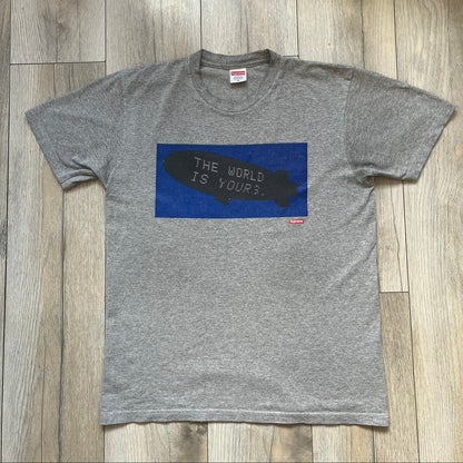 Supreme FW17 Scarface Blimp Gray Tee is a grey T-shirt from the Fall/Winter 2017 collection featuring a large front print from the film Scarface. The graphic shows the blimp scene with “The World Is Yours” text in black on a blue background, with a small red Supreme box logo printed below. Released in October 2017 as part of the Supreme x Scarface collaboration.
