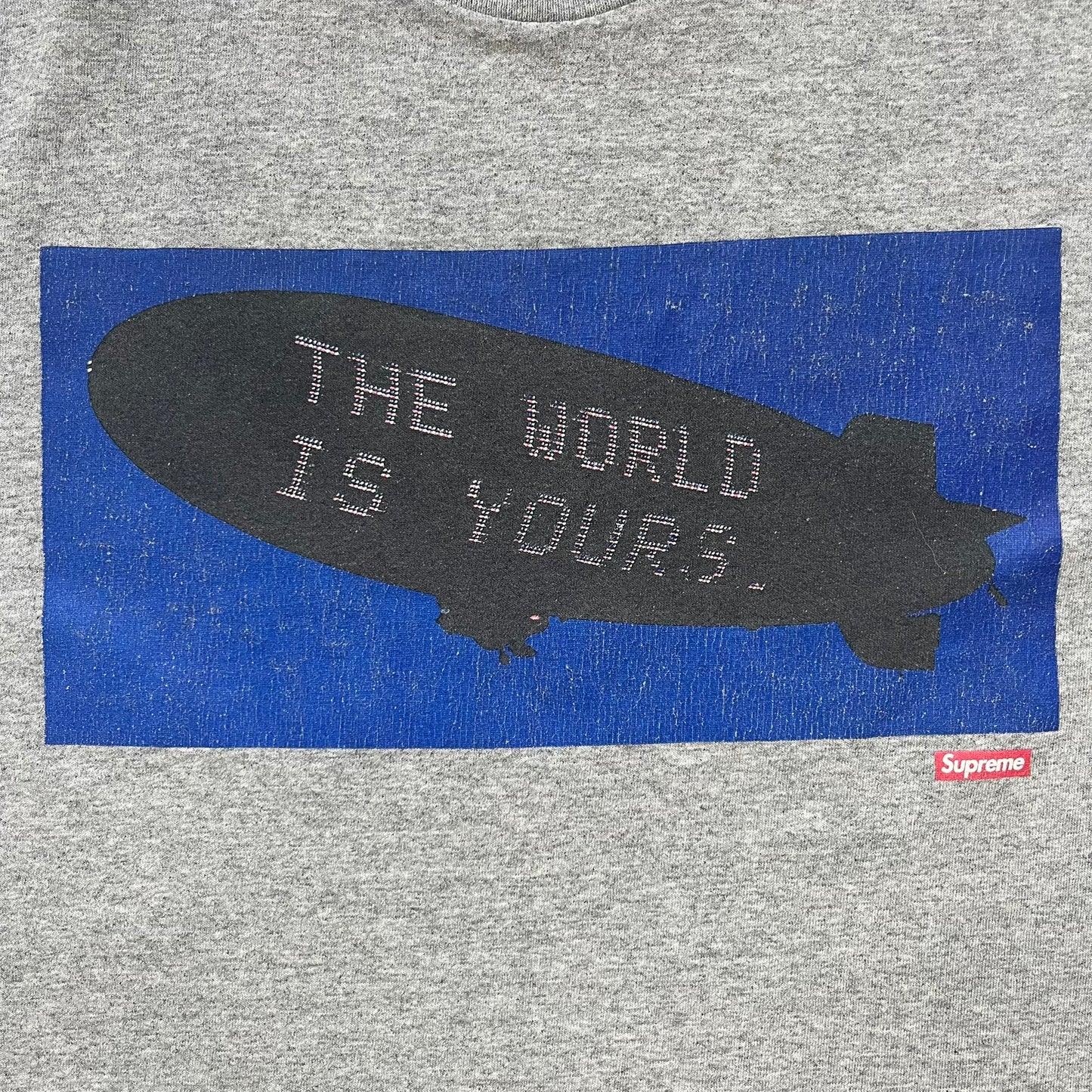 Supreme FW17 Scarface Blimp Gray Tee is a grey T-shirt from the Fall/Winter 2017 collection featuring a large front print from the film Scarface. The graphic shows the blimp scene with “The World Is Yours” text in black on a blue background, with a small red Supreme box logo printed below. Released in October 2017 as part of the Supreme x Scarface collaboration.