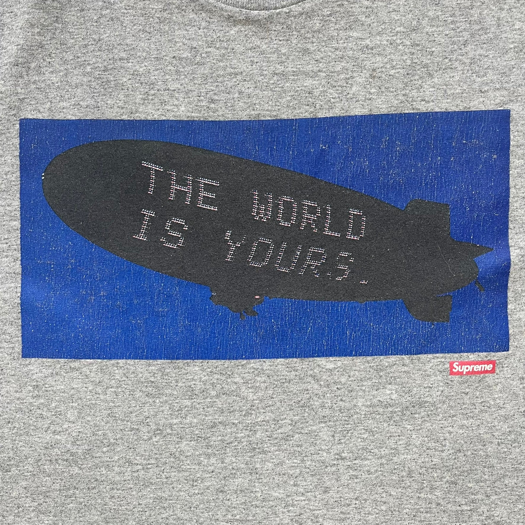 Supreme FW17 Scarface Blimp Gray Tee is a grey T-shirt from the Fall/Winter 2017 collection featuring a large front print from the film Scarface. The graphic shows the blimp scene with “The World Is Yours” text in black on a blue background, with a small red Supreme box logo printed below. Released in October 2017 as part of the Supreme x Scarface collaboration.