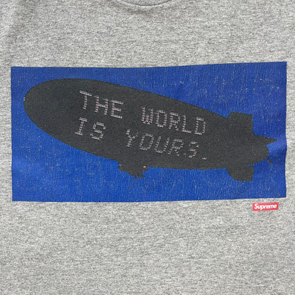 Supreme FW17 Scarface Blimp Gray Tee is a grey T-shirt from the Fall/Winter 2017 collection featuring a large front print from the film Scarface. The graphic shows the blimp scene with “The World Is Yours” text in black on a blue background, with a small red Supreme box logo printed below. Released in October 2017 as part of the Supreme x Scarface collaboration.