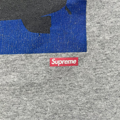 Supreme FW17 Scarface Blimp Gray Tee is a grey T-shirt from the Fall/Winter 2017 collection featuring a large front print from the film Scarface. The graphic shows the blimp scene with “The World Is Yours” text in black on a blue background, with a small red Supreme box logo printed below. Released in October 2017 as part of the Supreme x Scarface collaboration.