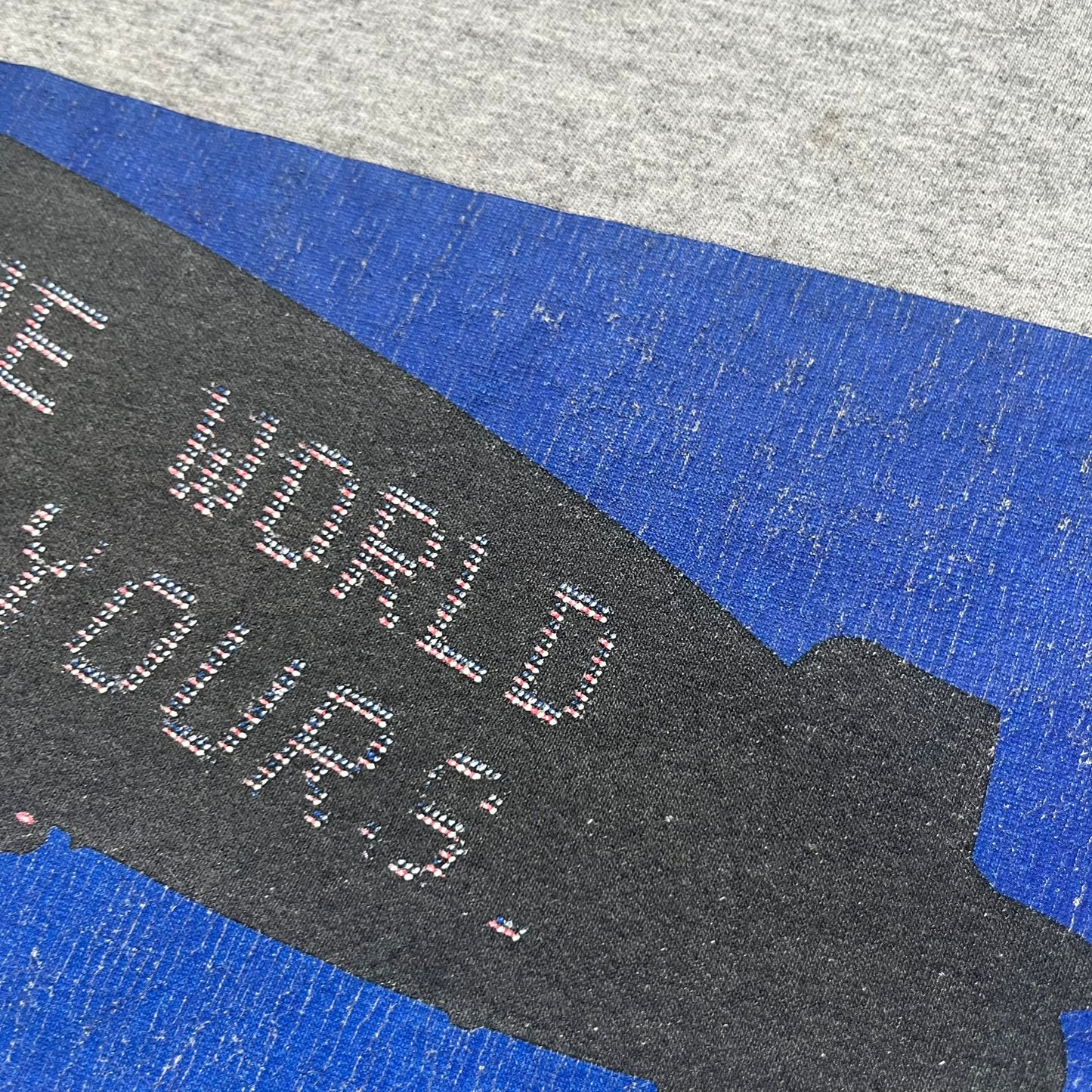 Supreme FW17 Scarface Blimp Gray Tee is a grey T-shirt from the Fall/Winter 2017 collection featuring a large front print from the film Scarface. The graphic shows the blimp scene with “The World Is Yours” text in black on a blue background, with a small red Supreme box logo printed below. Released in October 2017 as part of the Supreme x Scarface collaboration.