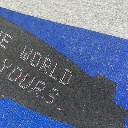 Supreme FW17 Scarface Blimp Gray Tee is a grey T-shirt from the Fall/Winter 2017 collection featuring a large front print from the film Scarface. The graphic shows the blimp scene with “The World Is Yours” text in black on a blue background, with a small red Supreme box logo printed below. Released in October 2017 as part of the Supreme x Scarface collaboration.