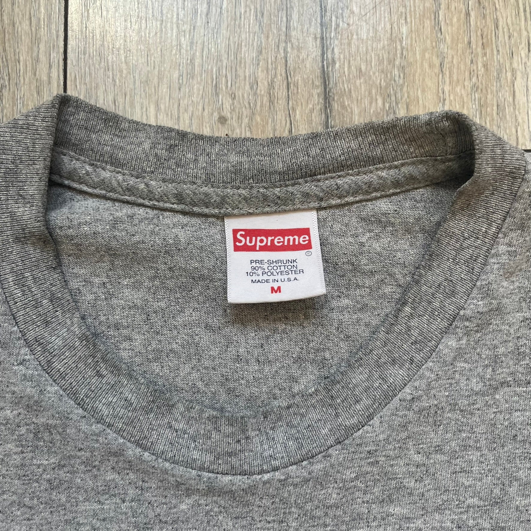 Supreme FW17 Scarface Blimp Gray Tee is a grey T-shirt from the Fall/Winter 2017 collection featuring a large front print from the film Scarface. The graphic shows the blimp scene with “The World Is Yours” text in black on a blue background, with a small red Supreme box logo printed below. Released in October 2017 as part of the Supreme x Scarface collaboration.