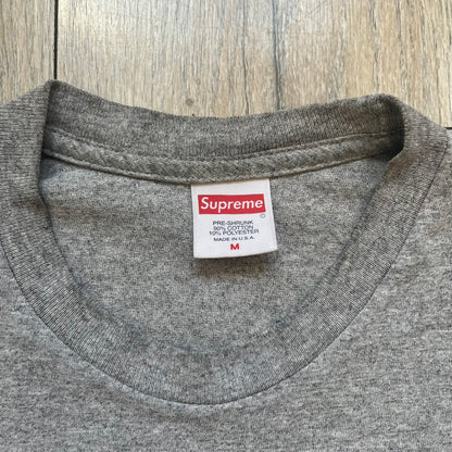 Supreme FW17 Scarface Blimp Gray Tee is a grey T-shirt from the Fall/Winter 2017 collection featuring a large front print from the film Scarface. The graphic shows the blimp scene with “The World Is Yours” text in black on a blue background, with a small red Supreme box logo printed below. Released in October 2017 as part of the Supreme x Scarface collaboration.