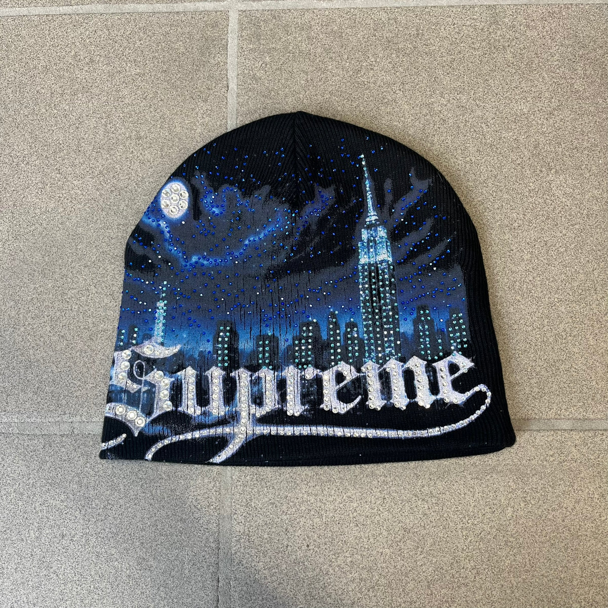 Supreme FW26 Cityscape Art Dealer Black Beanie is a black acrylic beanie with original artwork by Art Dealer, featuring a blue and white night city graphic with a moon and small stars across the sky on both sides. An old English-style Supreme logo sits on the front, and rhinestone appliqués in different sizes decorate the front and back. The print is stretchy, so any cracking effect that appears is just the print stretching with wear.