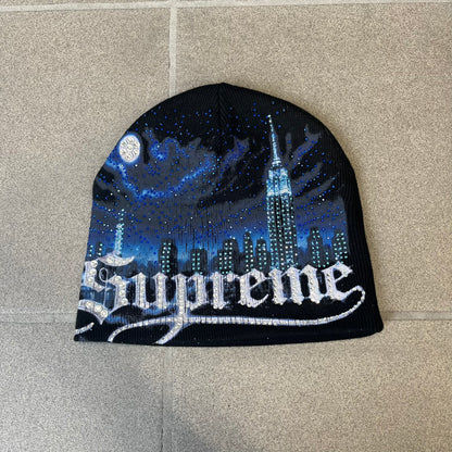 Supreme FW26 Cityscape Art Dealer Black Beanie is a black acrylic beanie with original artwork by Art Dealer, featuring a blue and white night city graphic with a moon and small stars across the sky on both sides. An old English-style Supreme logo sits on the front, and rhinestone appliqués in different sizes decorate the front and back. The print is stretchy, so any cracking effect that appears is just the print stretching with wear.