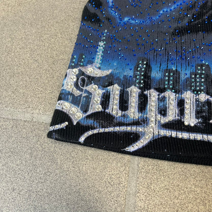 Supreme FW26 Cityscape Art Dealer Black Beanie is a black acrylic beanie with original artwork by Art Dealer, featuring a blue and white night city graphic with a moon and small stars across the sky on both sides. An old English-style Supreme logo sits on the front, and rhinestone appliqués in different sizes decorate the front and back. The print is stretchy, so any cracking effect that appears is just the print stretching with wear.