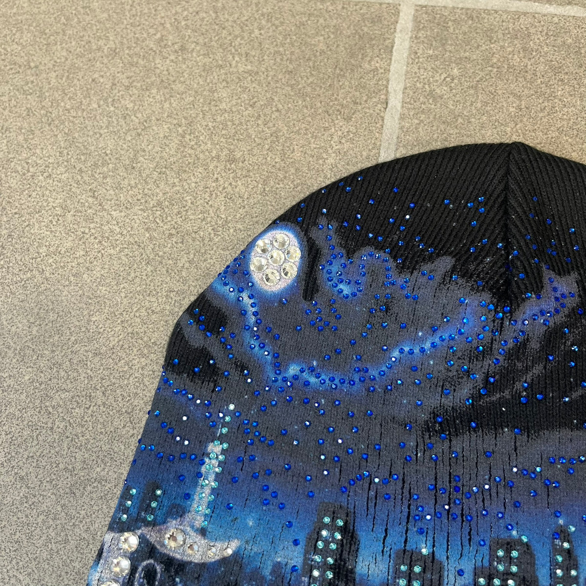 Supreme FW26 Cityscape Art Dealer Black Beanie is a black acrylic beanie with original artwork by Art Dealer, featuring a blue and white night city graphic with a moon and small stars across the sky on both sides. An old English-style Supreme logo sits on the front, and rhinestone appliqués in different sizes decorate the front and back. The print is stretchy, so any cracking effect that appears is just the print stretching with wear.