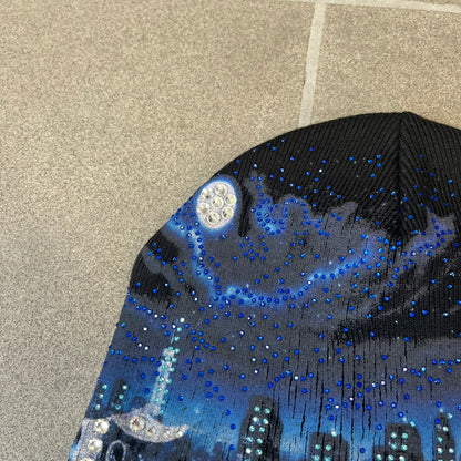 Supreme FW26 Cityscape Art Dealer Black Beanie is a black acrylic beanie with original artwork by Art Dealer, featuring a blue and white night city graphic with a moon and small stars across the sky on both sides. An old English-style Supreme logo sits on the front, and rhinestone appliqués in different sizes decorate the front and back. The print is stretchy, so any cracking effect that appears is just the print stretching with wear.