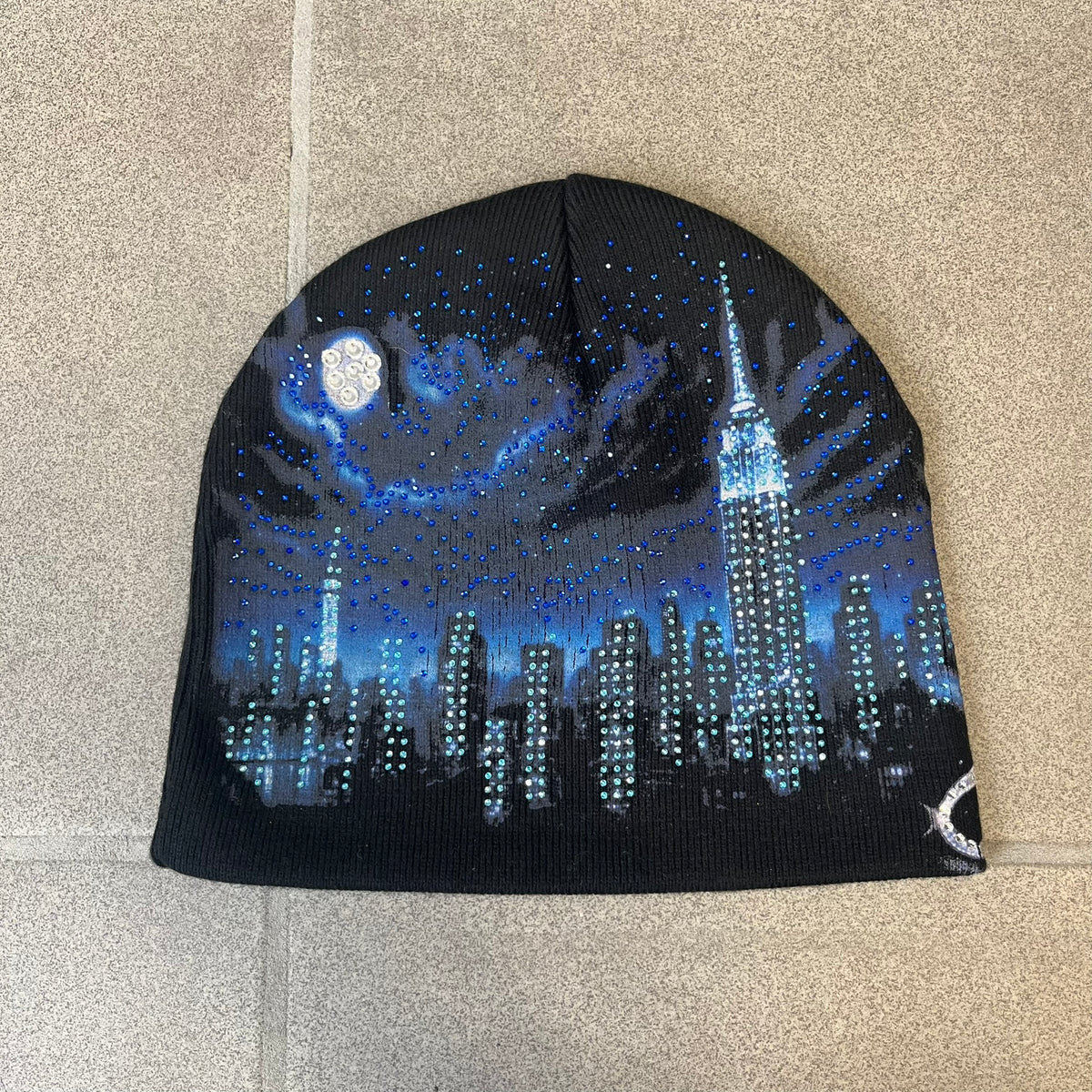 Supreme FW26 Cityscape Art Dealer Black Beanie is a black acrylic beanie with original artwork by Art Dealer, featuring a blue and white night city graphic with a moon and small stars across the sky on both sides. An old English-style Supreme logo sits on the front, and rhinestone appliqués in different sizes decorate the front and back. The print is stretchy, so any cracking effect that appears is just the print stretching with wear.